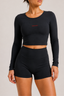 A woman modeling a black long-sleeve crop top and matching high-waisted black athletic shorts against a light gray studio background.