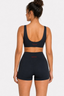A woman shown from the back wearing a black athletic set consisting of a scoop-neck sports bra and high-waisted shorts with small white 'PRINCE A DUTY' branding on the waistband and back of the bra, against a light gray studio background.