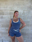 Stride Crop and Stride Shorts in Platinum, full front view of model with hand on hip against a grey brick wall, showing white piping and criss-cross waistband