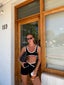 Stride Sports Bra in Black, lifestyle photo of model in sunglasses standing in a timber shop doorway, styled with matching Stride Shorts and holding an iced coffee
