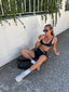 Stride Sports Bra and Stride Shorts in Black, lifestyle photo of model in sunglasses seated on a footpath sipping an iced drink, with white socks and sneakers