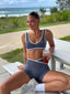 Stride Sports Bra and Stride Shorts in Platinum, lifestyle photo of smiling model seated at a beachside picnic table with an iced tea, ocean behind