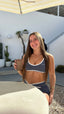 Stride Sports Bra in Platinum, lifestyle photo of smiling model seated at an outdoor café table holding an iced coffee, wearing the sports bra with white piping and matching Stride Shorts