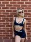 Stride Sports Bra and Stride Shorts in Black, lifestyle photo of smiling model in sunglasses leaning against a red brick wall, showing white piping and criss-cross waistband