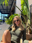 Daily Long Sleeve Top in Field Green, lifestyle photo of smiling model in sunglasses seated outdoors among tropical plants