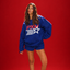 A woman modeling a royal blue PXM sweatshirt and matching shorts against a solid red studio background. The sweatshirt features a large red and white PXM logo with a star graphic on the front.