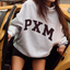 A woman wearing an oversized light gray sweatshirt with 'PXM' printed across the chest in dark brown block letters, sitting in the open door of a yellow car.