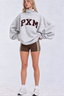 A woman modeling an oversized light gray sweatshirt with 'PXM' printed in dark brown across the chest, paired with brown athletic shorts featuring white side stripes, white socks, and white sneakers, against a light gray studio background.